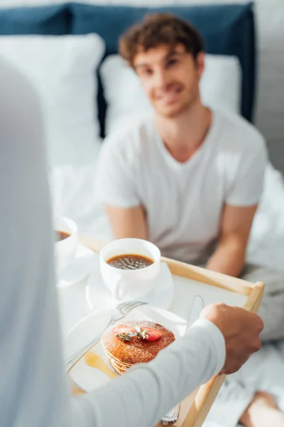 Selective focus of woman holding breakfast tray with coffee and pancakes near boyfriend in bedroom — Stock Photo