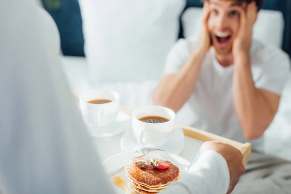 Selektiver Fokus der Frau, die Frühstück und Kaffee auf Tablett in der Nähe ihres aufgeregten Freundes im Schlafzimmer hält — Stockfoto