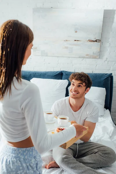 Enfoque selectivo del joven mirando a su novia con bandeja de desayuno en el dormitorio por la mañana - foto de stock