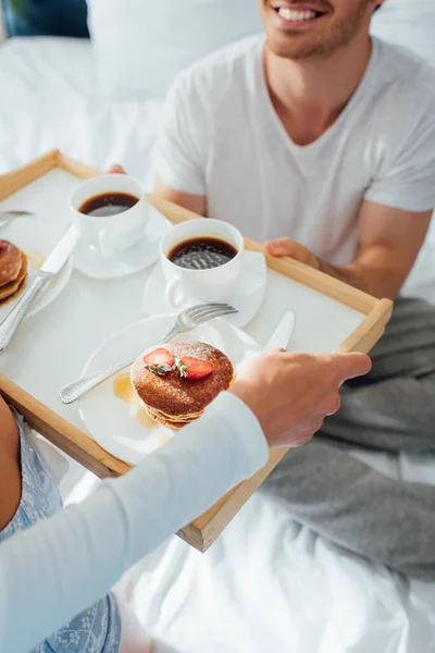 Cropped view of young woman holding pancakes and coffee on breakfast tray near boyfriend in pajamas on bed — Stock Photo