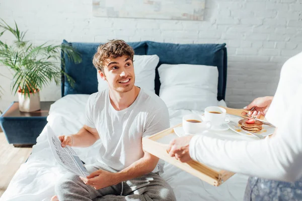 Selective focus of man with newspaper looking at girlfriend holding breakfast on tray in bedroom — Stock Photo