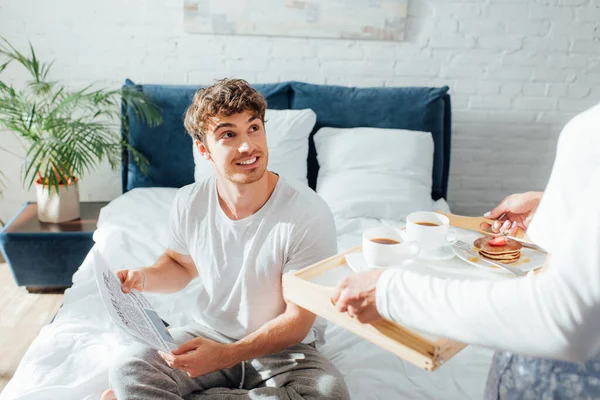 Concentration sélective de l'homme avec journal regardant petite amie tenant le petit déjeuner sur le plateau dans la chambre — Photo de stock