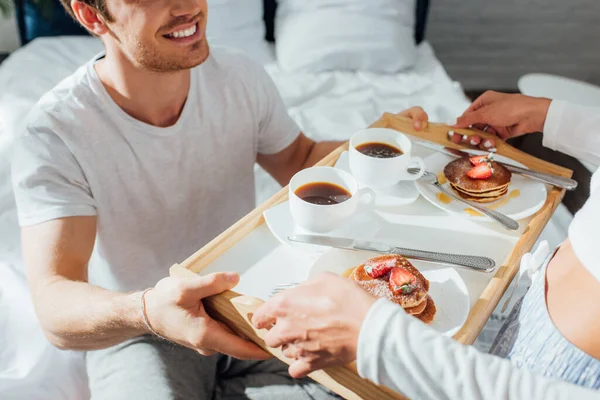 Cropped view of young couple in pajamas holding breakfast tray with pancakes and coffee on bed — Stock Photo