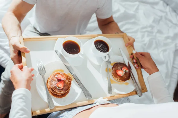 Vista de ángulo alto de la mujer sosteniendo panqueques y tazas de café en la bandeja del desayuno cerca de novio en la cama - foto de stock