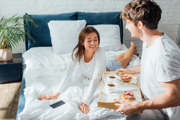 Foco seletivo da mulher de pijama olhando para o café da manhã na bandeja perto do namorado no quarto — Fotografia de Stock