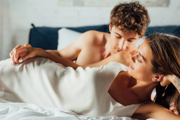 Homme musclé embrassant épaule et main touchante de femme séduisante en chemise de nuit sur le lit — Photo de stock