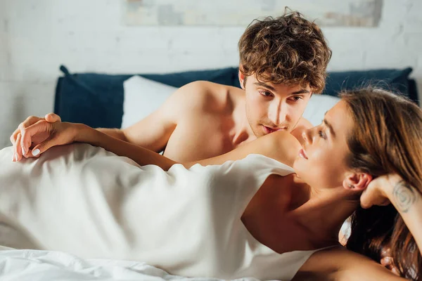 М'язистий чоловік цілує плече молодої дівчини в шовковій сукні на ліжку — стокове фото