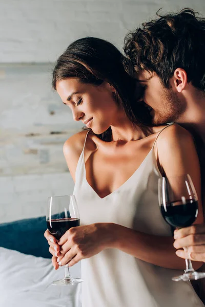 Selective focus of man kissing neck of girlfriend with glass of wine in bedroom — Stock Photo