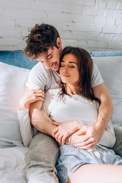 Selective focus of young man hugging girlfriend in pajamas on bed — Stock Photo