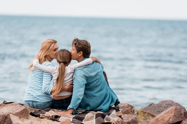 Back view of parents embracing and kissing daughter on plaid near sea — Stock Photo