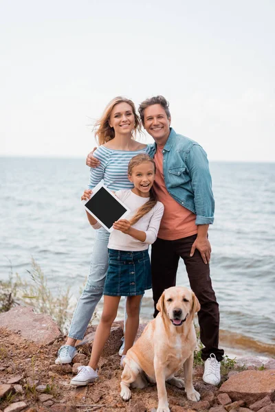 Child holding digital tablet near parents and golden retriever on beach near sea — Stock Photo