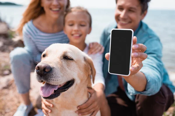 Selective focus of man holding smartphone near family and golden retriever on beach — Stock Photo