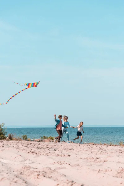 Foco seletivo do homem segurando pipa enquanto corre perto da família na praia — Fotografia de Stock