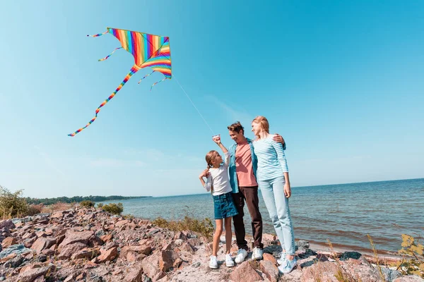 Mann umarmt Ehefrau und sieht Tochter mit Drachen am Strand an — Stockfoto