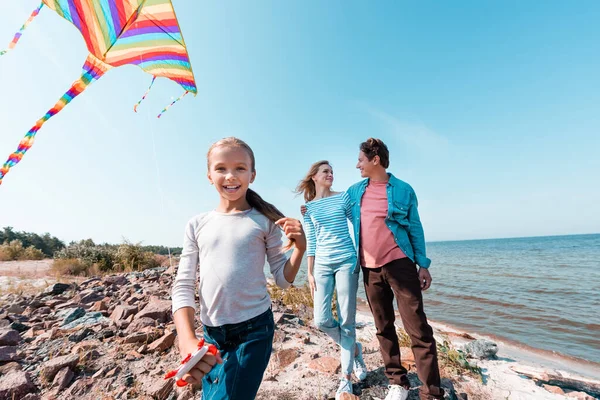 Selektiver Fokus eines Mädchens mit Drachen, das in die Kamera blickt, während die Eltern in der Nähe des Meeres stehen — Stockfoto