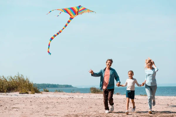 Familia con hija y cometa corriendo en la playa durante las vacaciones - foto de stock