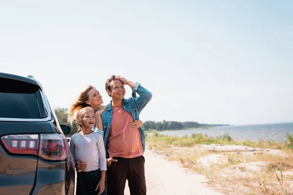 Enfoque selectivo de la mujer abrazando marido e hija emocionada cerca del coche en la playa - foto de stock