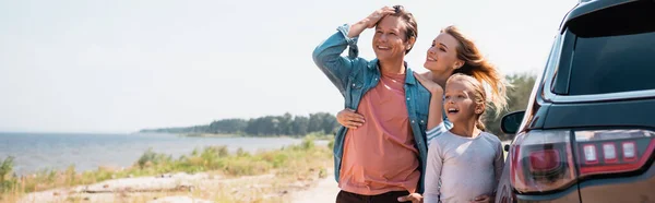 Panoramic shot of family with daughter hugging near car on beach — Stock Photo