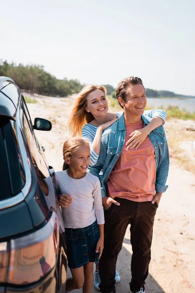 Enfoque selectivo de la mujer abrazando marido cerca de hija y auto en la playa - foto de stock