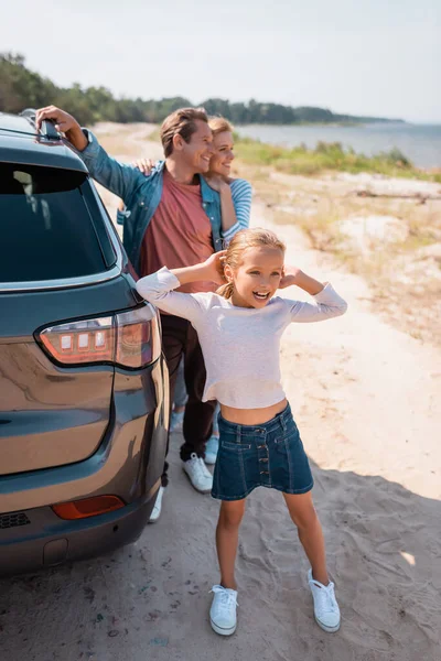 Foco seletivo da criança olhando para longe, enquanto os pais abraçando perto do carro na praia — Fotografia de Stock