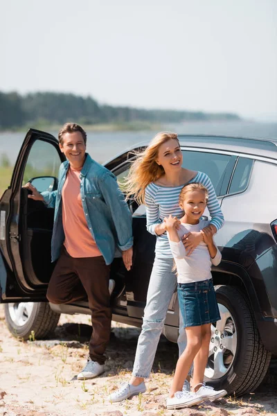 Selective focus of woman hugging child near man standing beside car on beach — Stock Photo