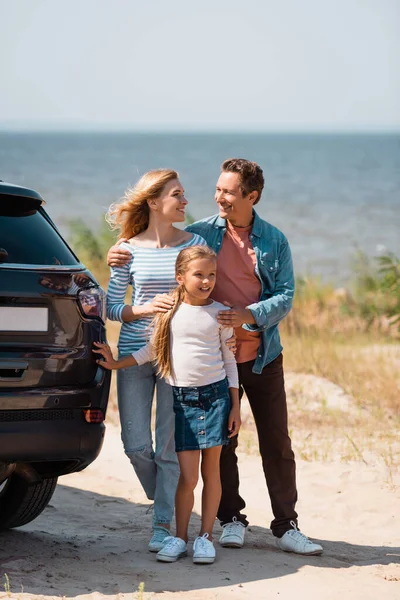 Избранное внимание мужчины и женщины, обнимающих дочь возле машины на берегу моря — стоковое фото
