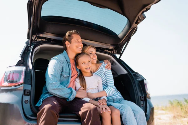 Семья смотрит в сторону, обнимаясь в багажнике машины в выходные — стоковое фото