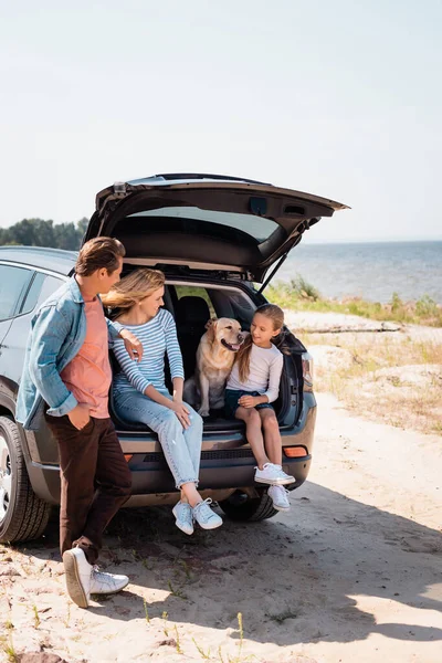 Selective focus of parents looking at daughter with golden retriever in car trunk on beach — Stock Photo