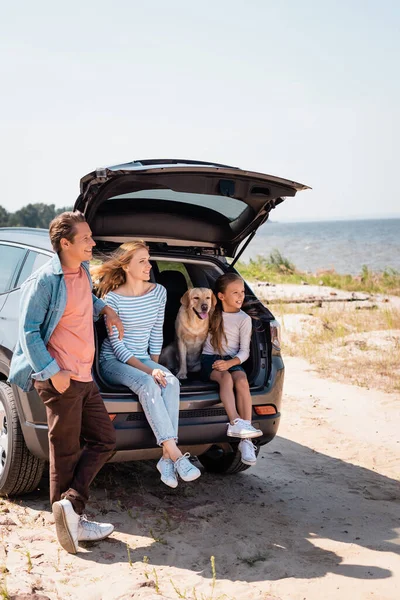 Selective focus of family with golden retriever looking away near auto on beach — Stock Photo