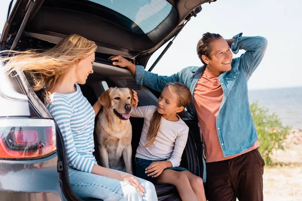 Selective focus of woman and child petting golden retriever in car trunk near father on beach — Stock Photo