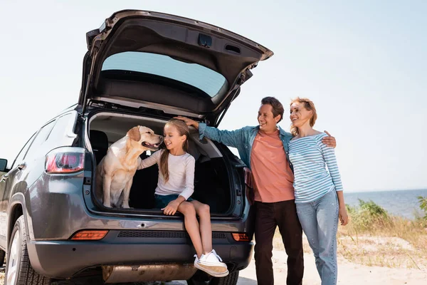 Homme étreignant femme près de fille et golden retriever dans un camion de voiture sur la plage — Photo de stock