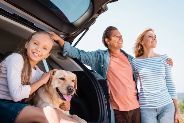 Enfoque selectivo de niña acariciando golden retriever cerca de los padres abrazando al lado del coche al aire libre - foto de stock