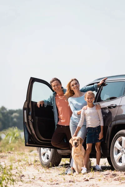 Selective focus of family with golden retriever looking at camera while standing near car outdoors — Stock Photo