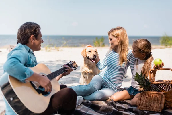 Focus selettivo di donna e bambino che giocano con golden retriever vicino al padre con chitarra acustica sulla spiaggia — Foto stock