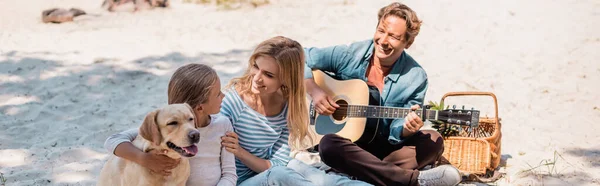 Colpo panoramico di donna che abbraccia bambino vicino golden retriever e marito con chitarra acustica sulla spiaggia — Foto stock