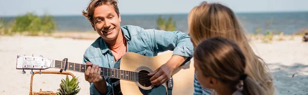Imagem horizontal do homem tocando guitarra acústica perto da família na praia — Fotografia de Stock