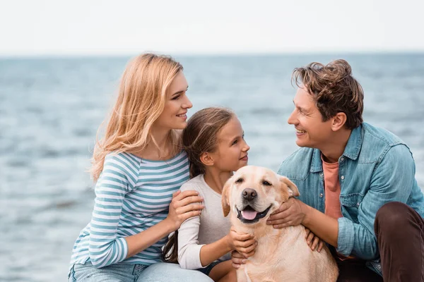 Pareja con hija y golden retriever mirándose en la playa - foto de stock