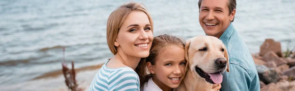 Foto panorâmica de pais e filha com golden retriever olhando para a câmera na praia — Fotografia de Stock