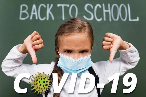 Selective focus of displeased schoolgirl in medical mask showing thumbs down near chalkboard with inscription and covid-19 lettering in classroom — Stock Photo