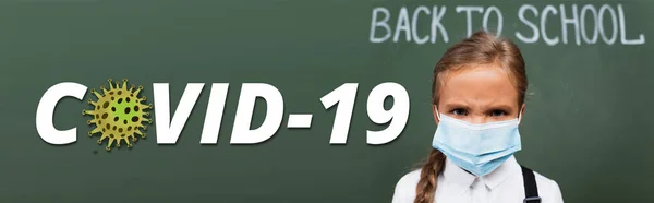 Horizontal image of displeased schoolgirl in medical mask near chalkboard and covid-19 lettering in classroom — Stock Photo