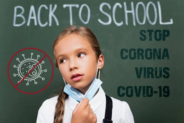 Enfoque selectivo de colegiala agotada quitándose la máscara protectora cerca de pizarra, corona de parada, virus, letras covid-19 y la ilustración - foto de stock