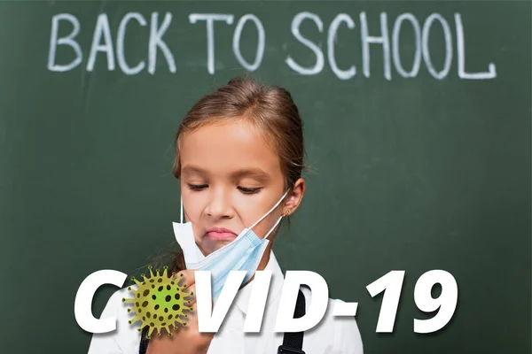 Selective focus of displeased schoolgirl touching protective mask near chalkboard and covid-19 lettering in classroom — Stock Photo