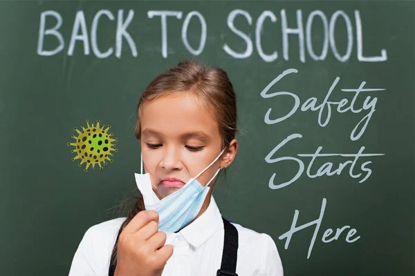 Foyer sélectif de l'écolière mécontente toucher masque de protection près de tableau noir avec la sécurité commence ici lettrage en classe — Photo de stock