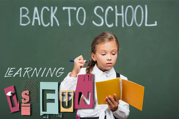 Concentrated schoolgirl holding copy book and pen near chalkboard and learning is fun lettering in classroom — Stock Photo