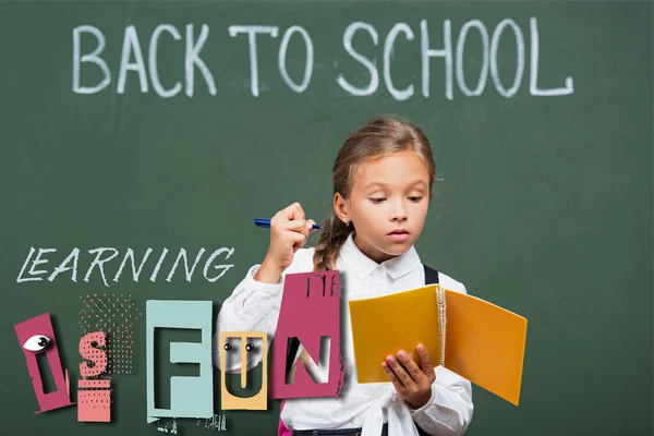 Concentrated schoolgirl holding copy book and pen near chalkboard and learning is fun lettering in classroom — Stock Photo