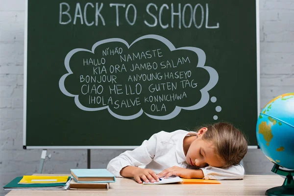 Exhausted schoolgirl sleeping at desk near globe and chalkboard with back to school and greeting lettering — Stock Photo