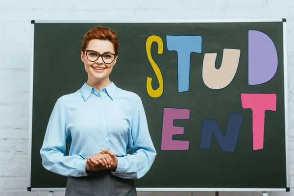 Enseignant en lunettes regardant la caméra tout en se tenant les mains serrées près du tableau avec lettrage étudiant — Photo de stock