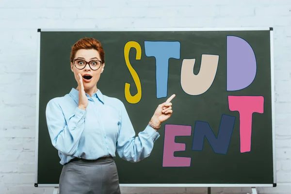 Impactado maestro tocando la cara y señalando con el dedo en pizarra con letras de estudiante - foto de stock