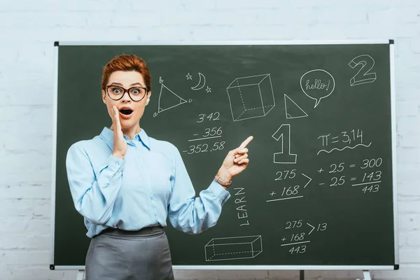 Shocked geometry teacher touching face and pointing with finger at chalkboard with illustration — Stock Photo