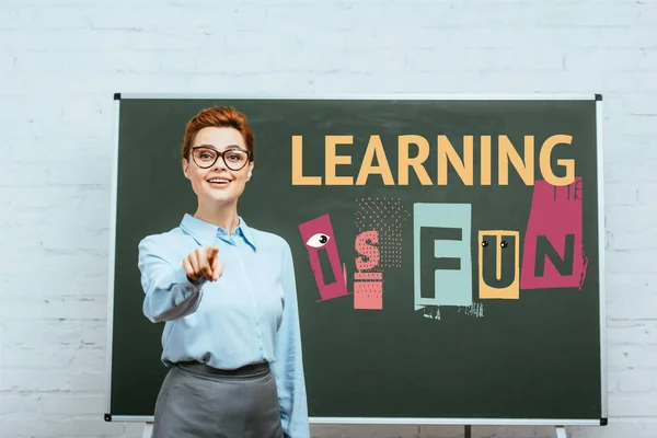 Teacher looking at camera and pointing with finger near chalkboard with learning is fun lettering — Stock Photo